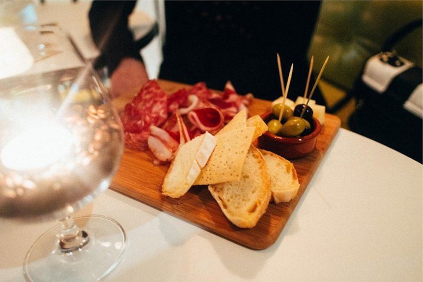 A wooden board with sliced bread, cheese, olives, and meatsticks. 2