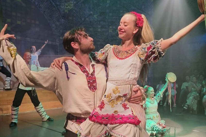 A stage with performers in traditional costumes, a man in a white shirt with red embroidery, and a woman in a pink dress with floral patterns. 3
