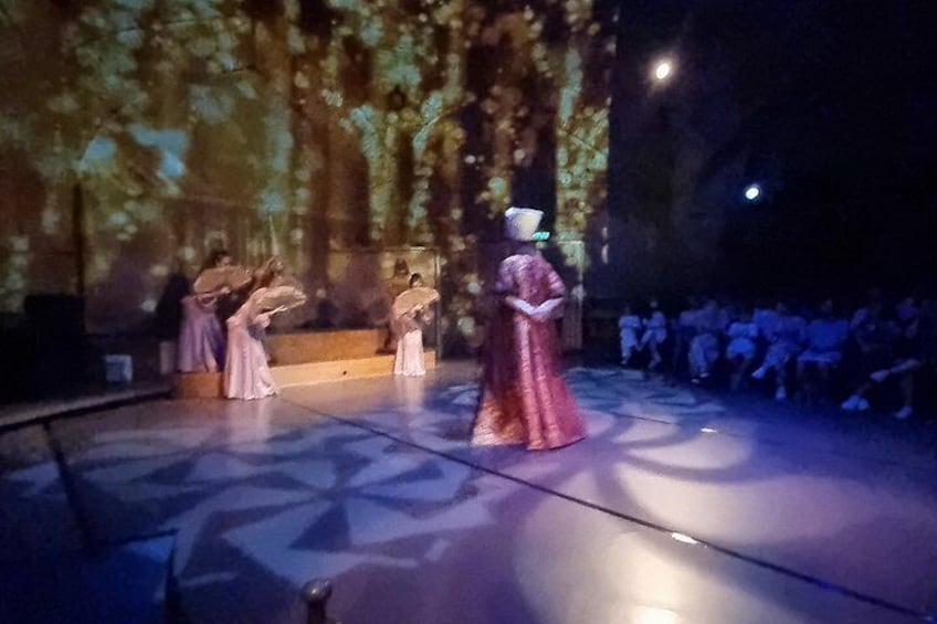 A stage with performers in period costumes, a large screen in the background, and an audience seated in rows. 5