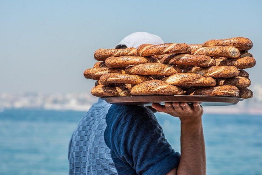 A person carrying a tray of bread over their head with a body of water and a distant city skyline in the background. 1