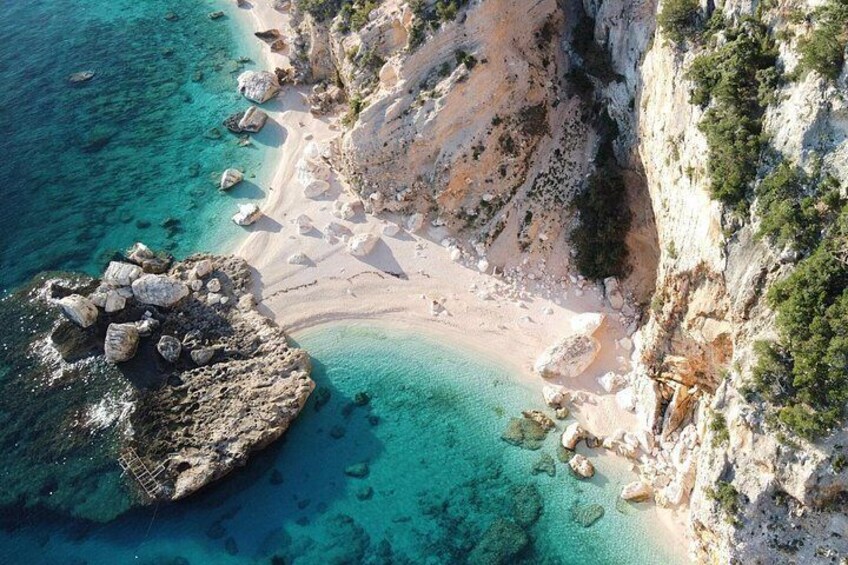 A coastal landscape with clear turquoise waters, a sandy beach, rocky outcrops, and sparse vegetation. 2