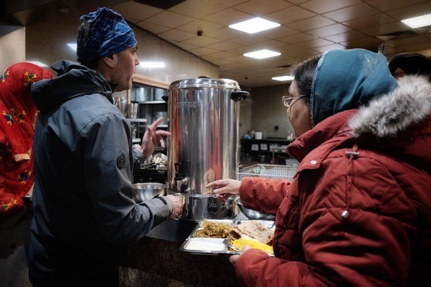 A person in a red jacket interacting with a service counter, another person in a blue jacket, and a large stainless steel container. 2