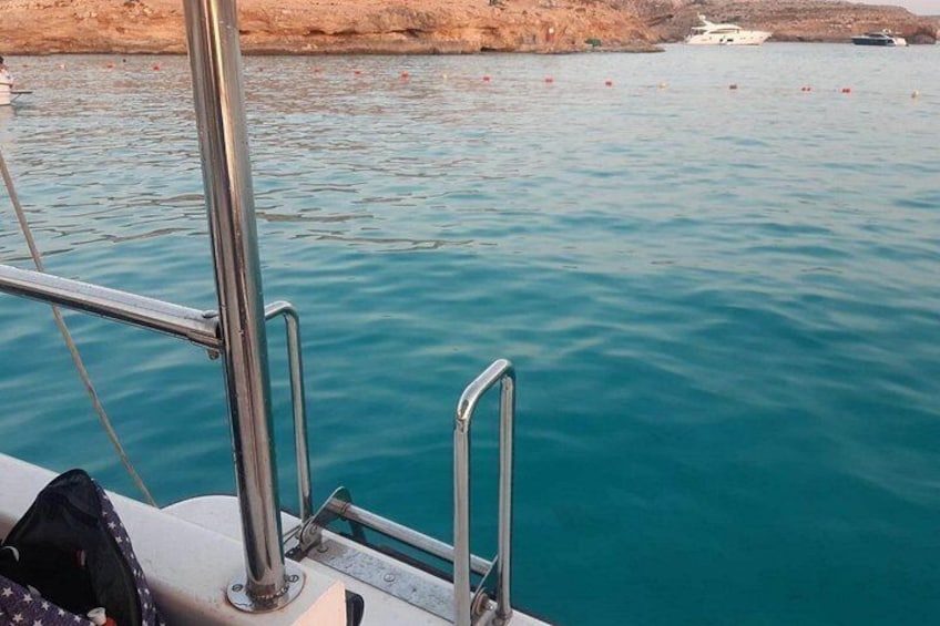 A boat's metal railing and part of its deck, with calm turquoise water and a rocky shoreline in the background. 1