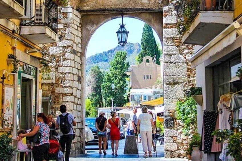 A stone archway with people walking through, shops on either side, and a mountain in the background. 4