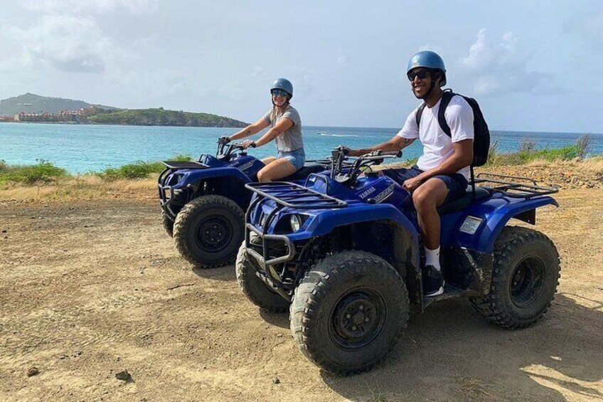 Dos personas en cuatrimotos azules sobre un sendero de tierra junto a un cuerpo de agua. 2