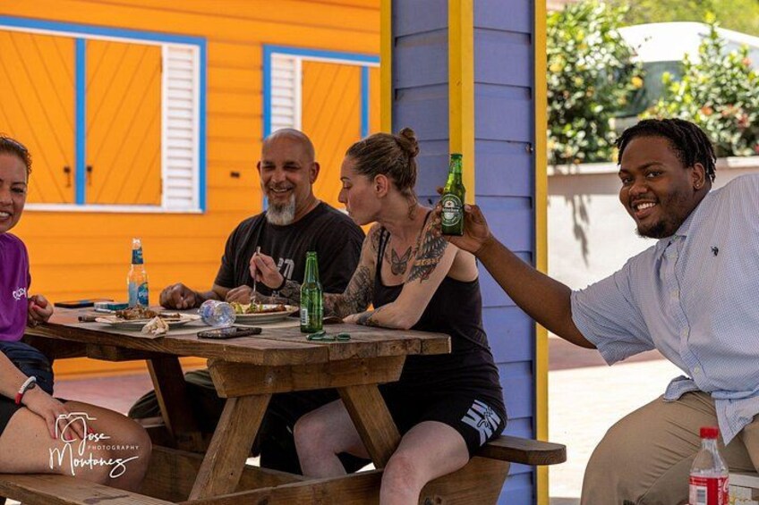 A group of people sitting at a wooden table outdoors, with a building in the background. 1