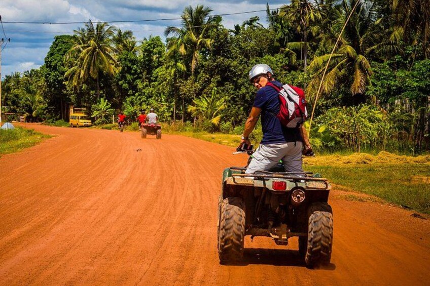 Une personne conduit un véhicule tout-terrain sur un chemin de terre, avec des palmiers et un véhicule jaune en arrière-plan. 1