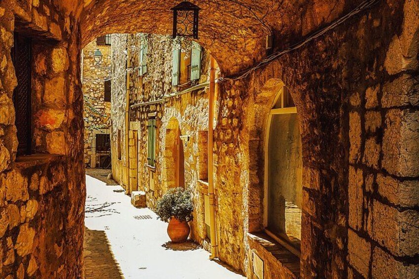 A narrow stone alleyway with arched doorways and windows, a potted plant, and a snow-covered ground. 2