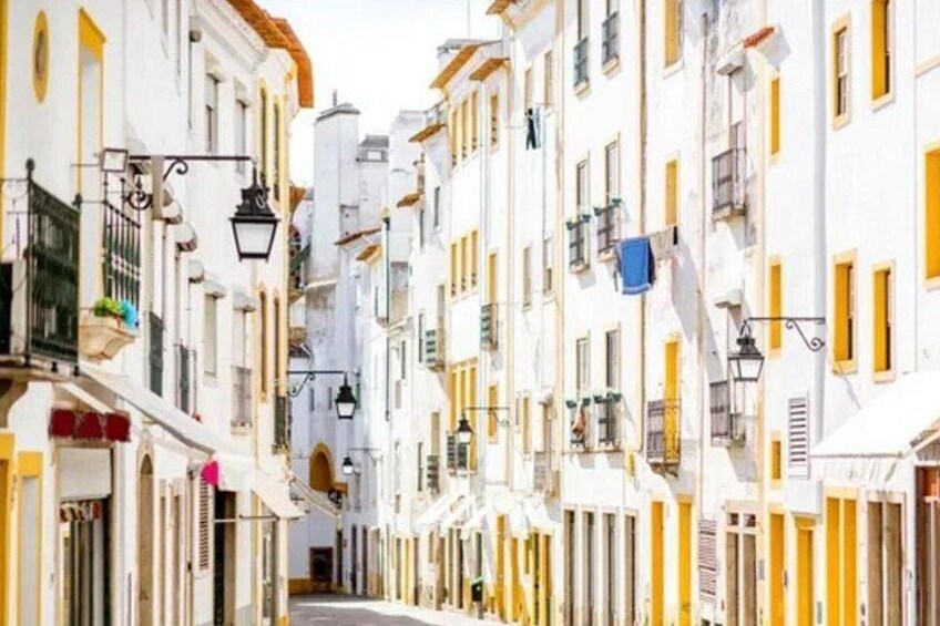 A narrow street lined with white buildings with yellow accents, featuring balconies and hanging laundry. 2