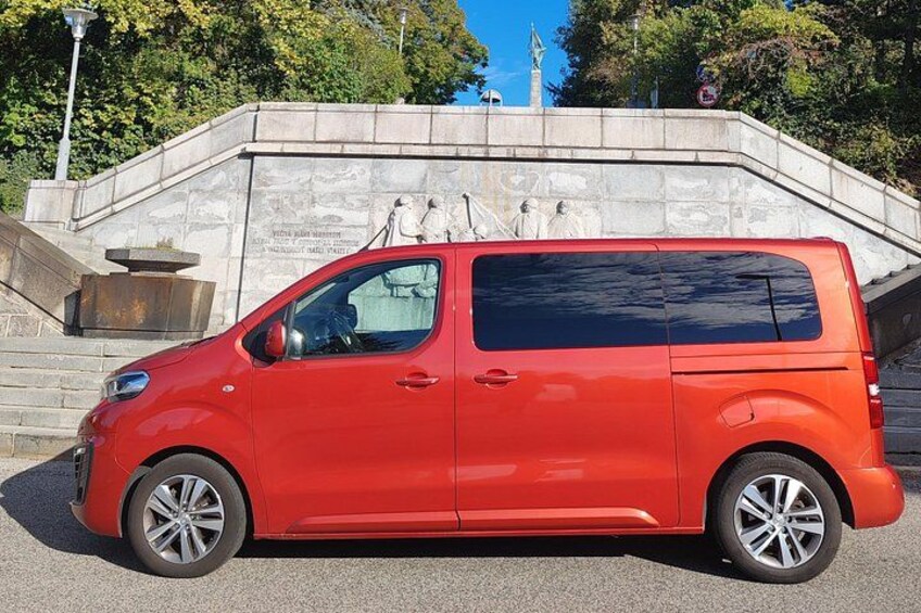 A red van parked on a paved area with a stone monument and a statue in the background. 3