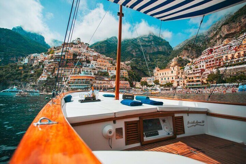 Un barco con cubierta de madera y cojines azules, navegando cerca de un pueblo costero con edificios de colores y montañas al fondo. 2