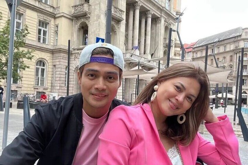 A person in a pink jacket and a cap, with a building featuring classical architecture in the background. 1