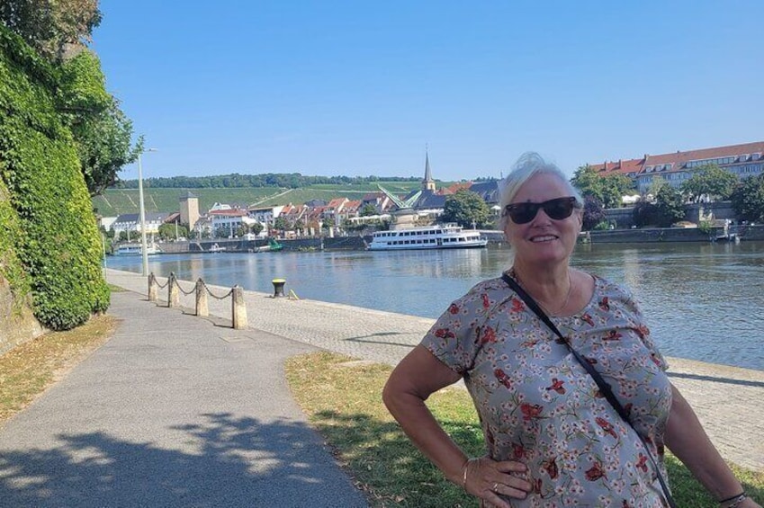 A woman standing by a riverside walkway with a boat in the background. 3