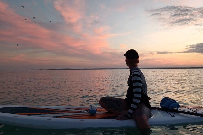 A person sitting on a paddleboard at sunset with a cup and a bag beside them. 2