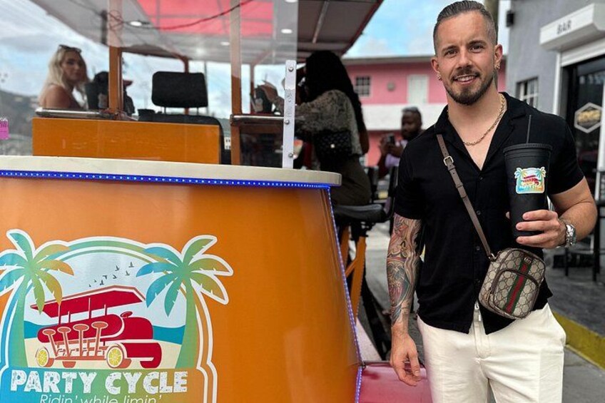 A man with tattoos holding a drink and a bag, standing in front of a colorful kiosk with a tropical design. 2