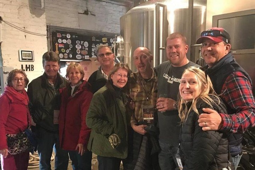 A group of people in a brewery setting with stainless steel tanks and a wall with various stickers. 2