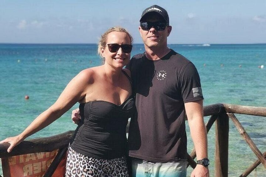 A couple standing on a wooden deck with a clear blue sea and sky in the background. 3