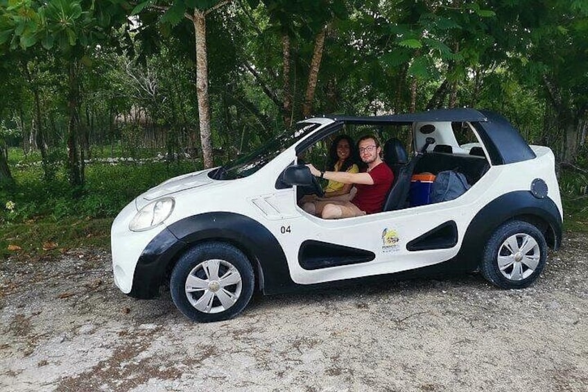 A white and black car with two passengers, parked on a gravel path surrounded by greenery. 1