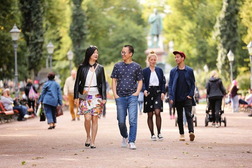 En gruppe mennesker går på en belagt sti i en park med træer og en statue i baggrunden. 5