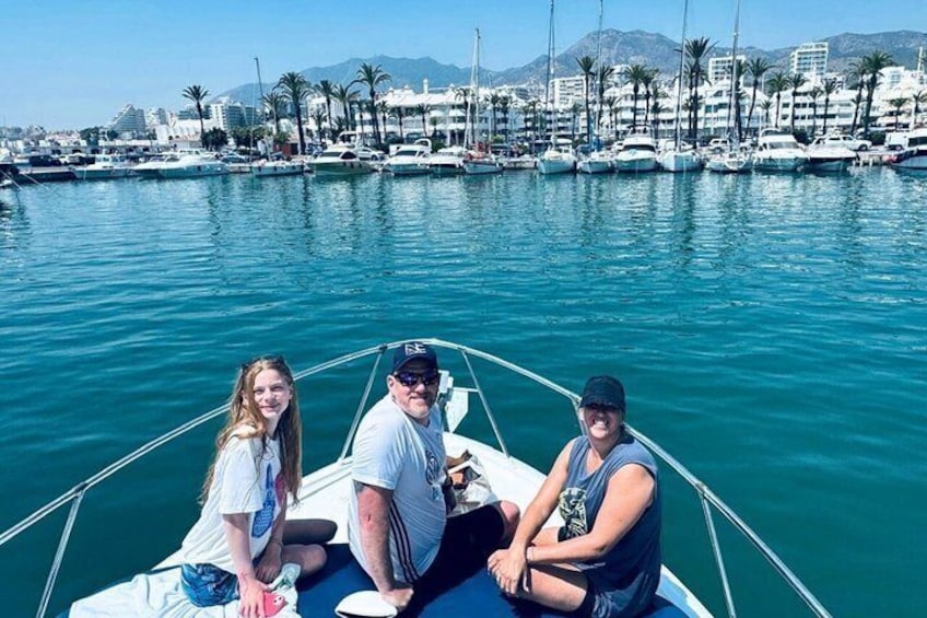 Trois personnes se trouvent sur un bateau dans une marina, avec des yachts et des bâtiments en arrière-plan. 5