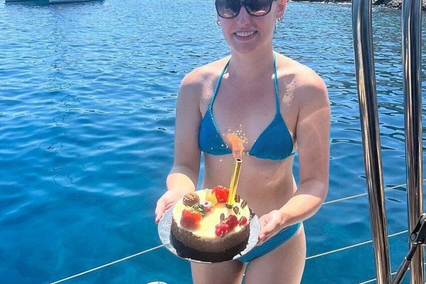 En person holder en kage med et tændt lys, iført en blå bikini, og står på en båd med vand i baggrunden. 4