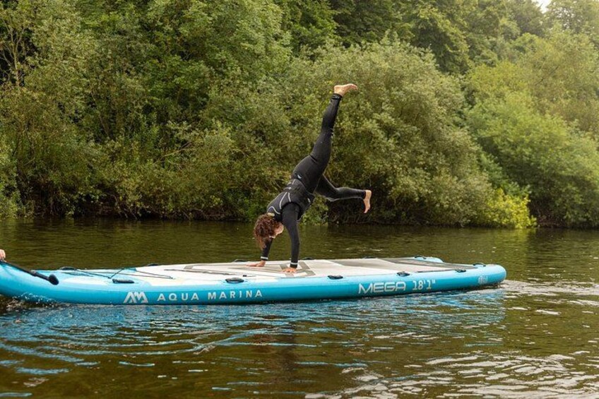 Une personne fait la verticale sur une planche gonflable bleue portant l’inscription « AQUA MARINA MEGA 181 », dans une rivière bordée de verdure. 2