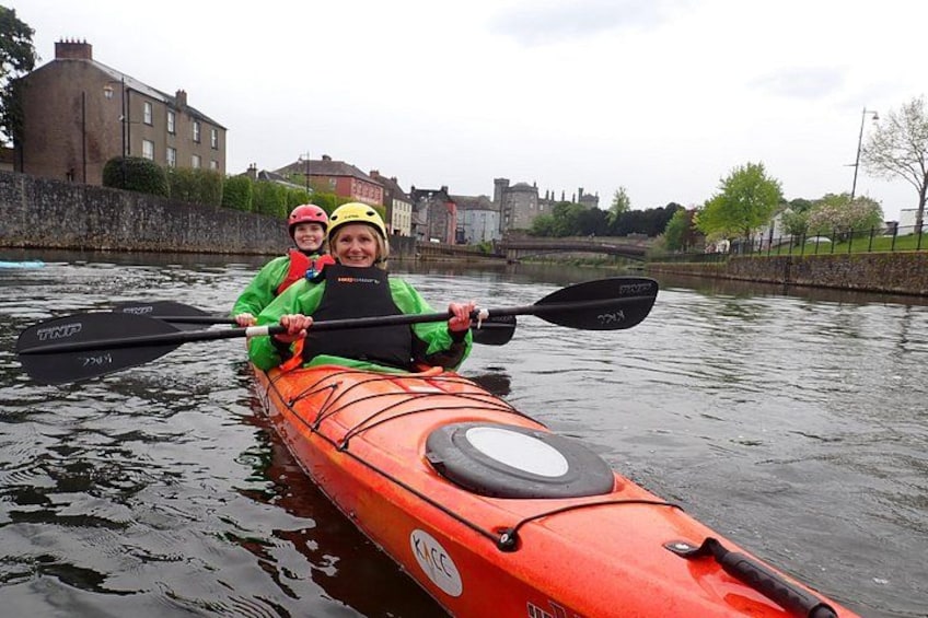 Deux personnes pratiquent le kayak sur une rivière, portant des casques et des gilets de sauvetage, avec des bâtiments et un pont en arrière-plan. 1
