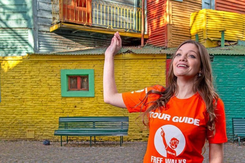 Una persona con una camiseta naranja que dice 'TOUR GUIDE' y 'FREE WILLOWS', de pie frente a un edificio amarillo con un ventanal verde y un banco. 4