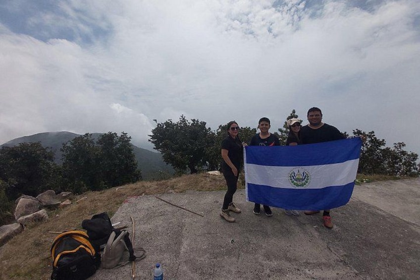 Un groupe de personnes tenant un drapeau, un sac à dos et une bouteille d’eau, se tenant sur une surface en béton, avec des arbres et des montagnes en arrière-plan. 5