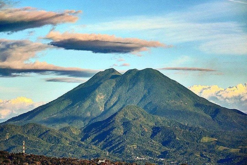 Une montagne avec un sommet, entourée de zones boisées et surplombée d’un ciel dégagé parsemé de nuages. 1