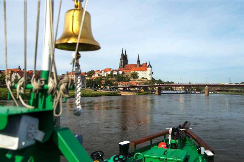 Un barco verde con una campana en primer plano, un río y un castillo con techos rojos al fondo. 1