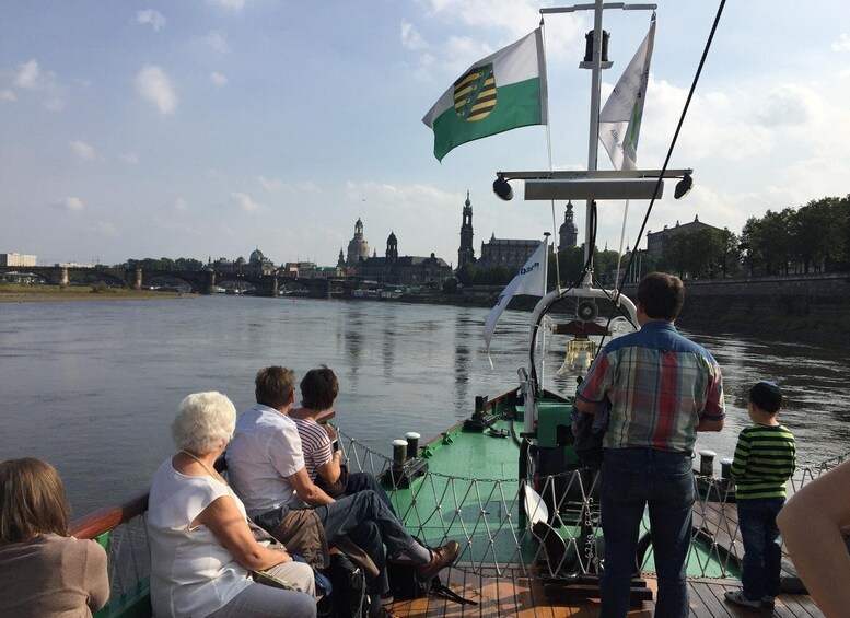 Un bote con pasajeros en un río, un paisaje urbano con edificios históricos al fondo y banderas en el bote. 4