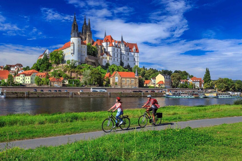 Dos ciclistas en un camino junto a un río, con un castillo histórico al fondo. 5