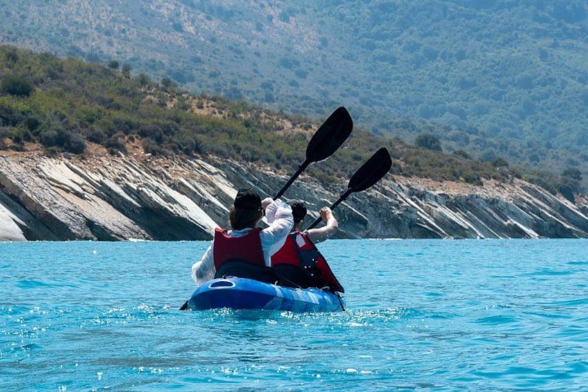 Two people kayaking on a calm body of water with a rocky shoreline and hills in the background. 3