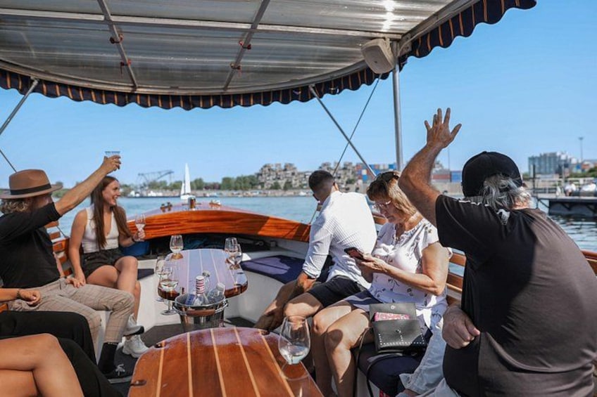 Un grupo de personas en un barco con una mesa, bebidas y un paisaje urbano al fondo. 2
