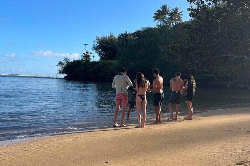 Een groep mensen staat op een zanderig strand bij het water, met bomen en een heldere lucht op de achtergrond. 2