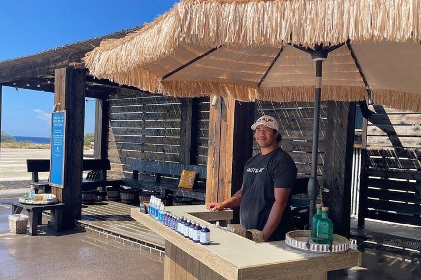 A man standing under a thatched umbrella at a beachside kiosk with a wooden structure, a signboard, and a display of bottled water. 1