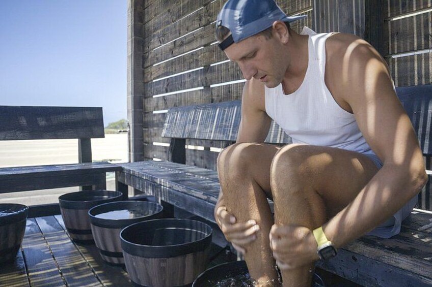 A person sitting on a wooden bench outdoors, with metal buckets and a wooden structure in the background. 5