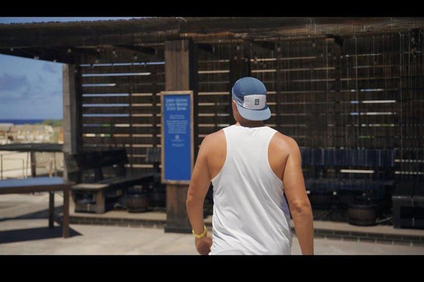 A person walking on a concrete path, wearing a white tank top and a blue cap, with a metal structure and a blue sign in the background. 2