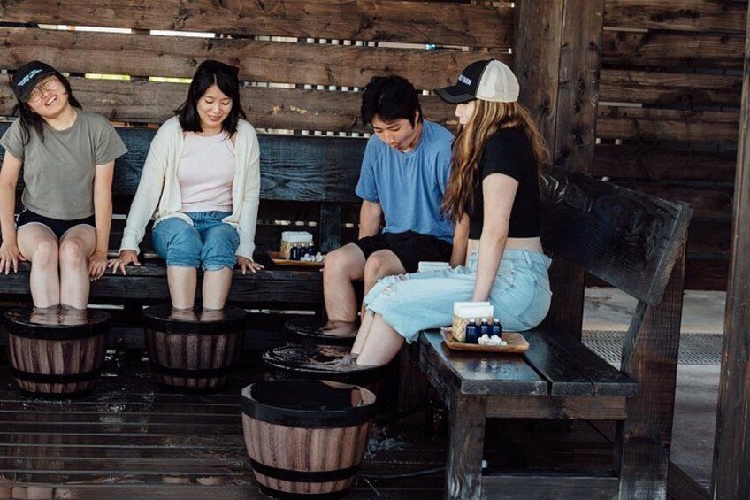 Four people sitting on wooden benches outdoors, with one person standing and holding a tray with bottles. 3