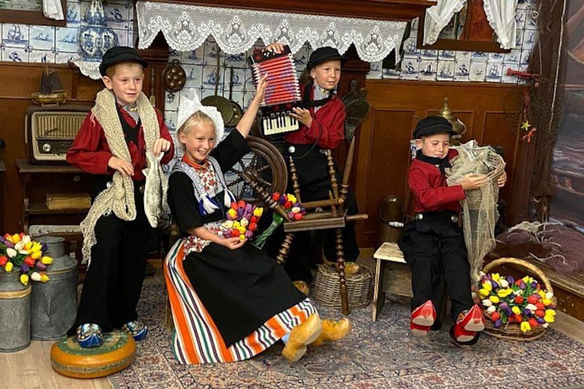 En traditionell miljö med en kvinna i en svart klänning och ett randigt förkläde, omgiven av barn i tidstypiska kostymer. 2