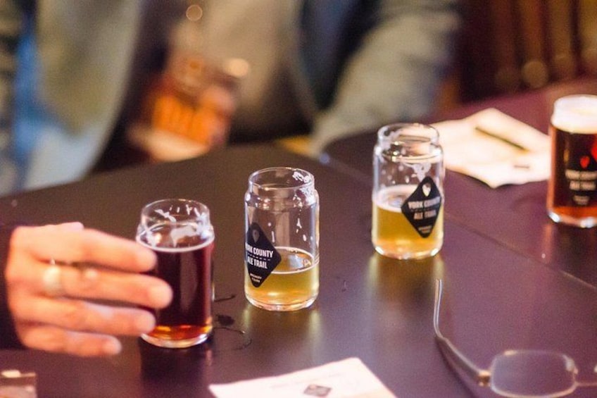 En person holder et glas øl, med tre andre glas på et bord, hvoraf det ene er mærket 'Vine County Trail'. 3