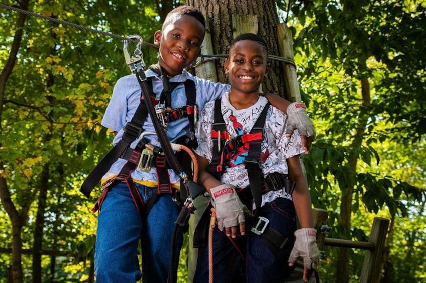 Two individuals wearing harnesses and safety gear, standing on a wooden platform in a forested area. 3