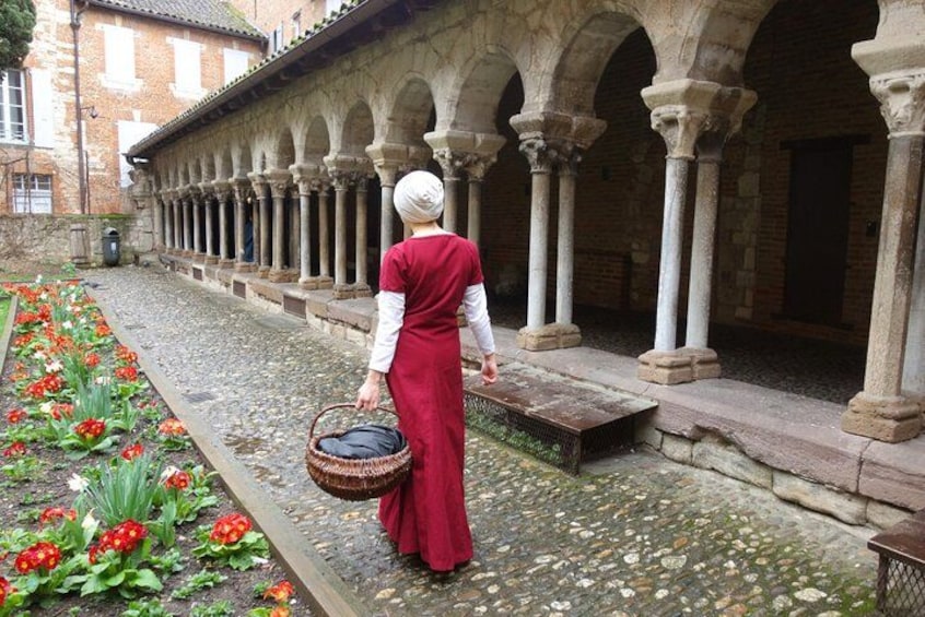 A person in a red dress walking along a cobblestone path with a basket, surrounded by a garden with red flowers and a building with arches. 3