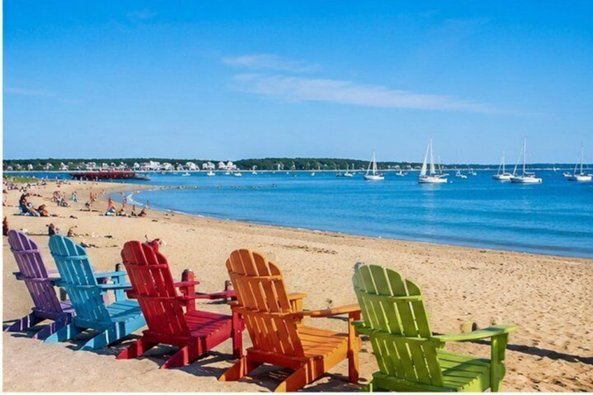 A beach with colorful Adirondack chairs and sailboats on the water. 5