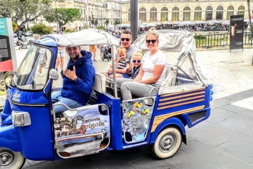 A blue tuk-tuk with stickers and a sign that reads "Antonia Pigola" driving on a city street. 3