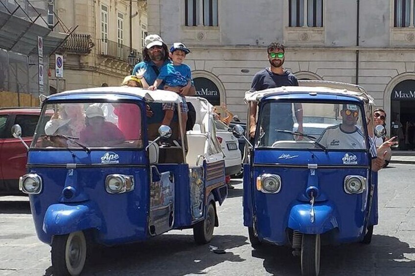 Two vintage blue tuk-tuks with passengers, parked on a street with buildings in the background. 2