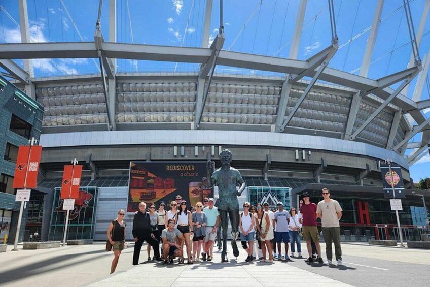 A group of people standing in front of a modern stadium with a large glass roof and a statue. 5