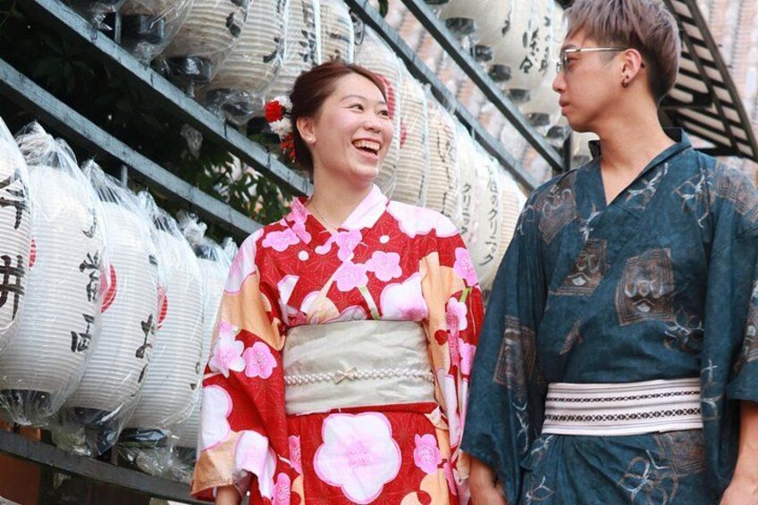 Two people in traditional Japanese attire, a woman in a red kimono with floral patterns and a man in a dark blue kimono with a geometric design, standing in front of a shelf with white ceramic items. 2