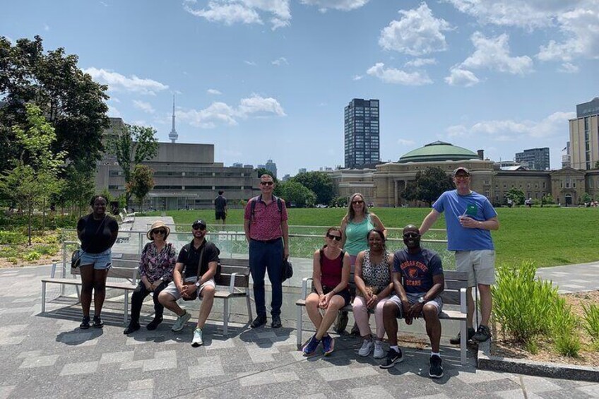 A group of people sitting and standing on a bench in a park with buildings and a tower in the background. 3